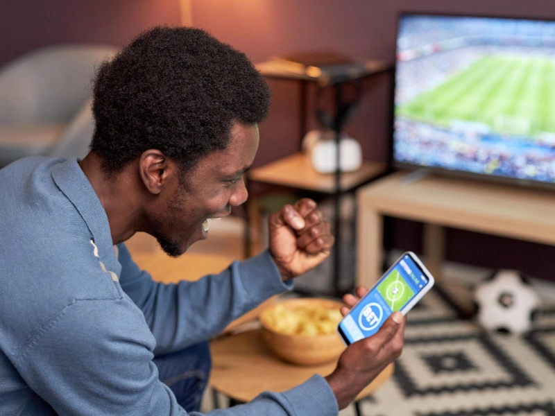 An excited man sitting on a couch watching a sports match on TV while holding a smartphone showing a cricket betting app.