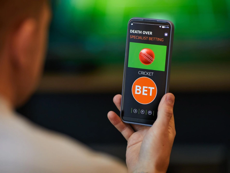 A man looking at a smartphone showing a cricket betting interface labeled Death Over Specialist Betting with a cricket ball and an orange BET button.